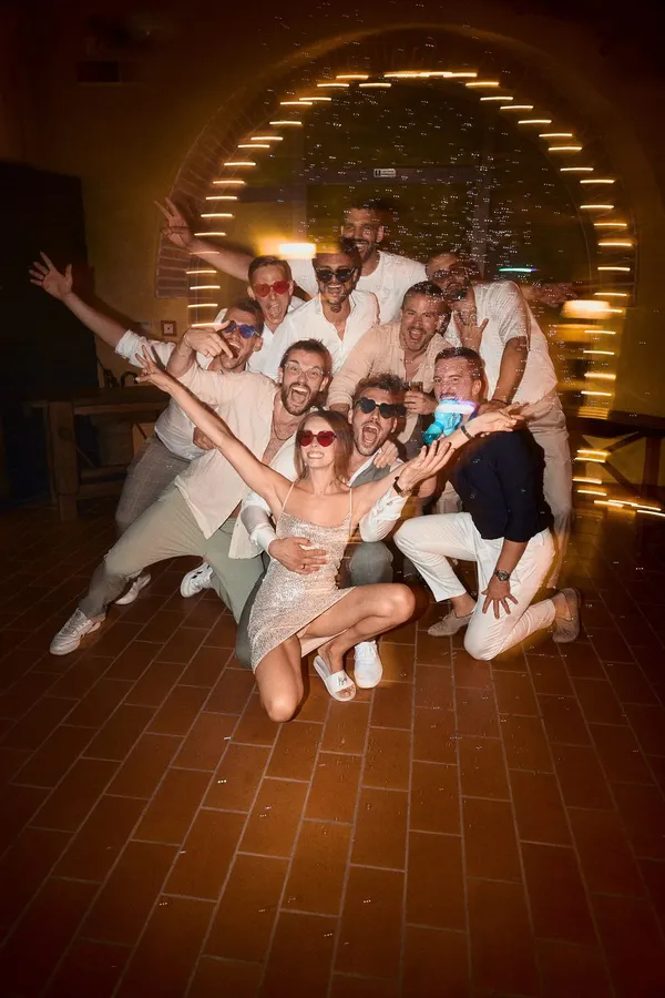 Exuberant group dressed in white poses beneath a lit archway on a tiled floor, flash by Kilian Amrehn