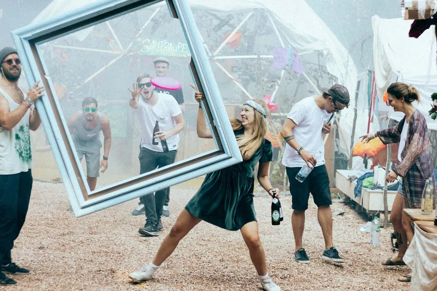 Revellers on a festival site, a woman carries a large pale blue picture frame, flash by Kilian Amrehn