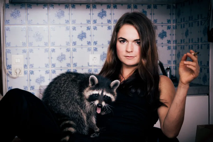 Woman with a cigarette and a raccoon on her lap in front of blue and white tiles, flash by Kilian Amrehn