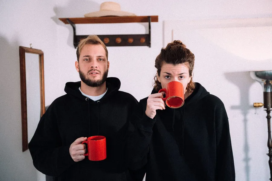 Couple in black hoodies holding red coffee mugs in a bright hallway, flash by Kilian Amrehn