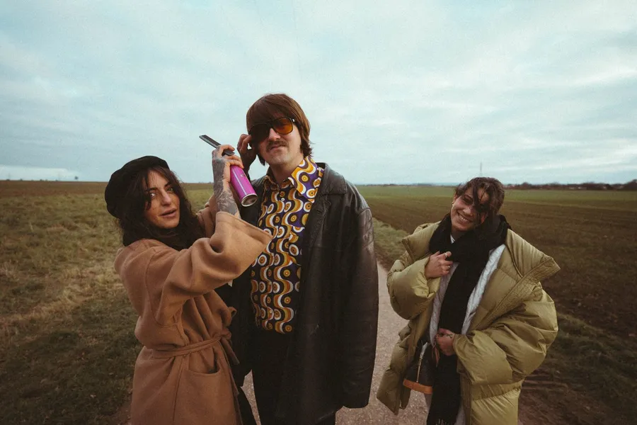 Three friends in coats and retro outfits on a field track under a grey sky, flash by Kilian Amrehn