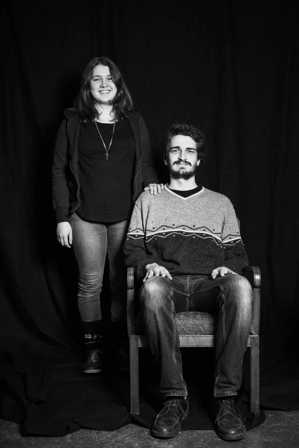 Couple before a black curtain, he sits on a chair, she stands beside him, black and white, flash by Kilian Amrehn