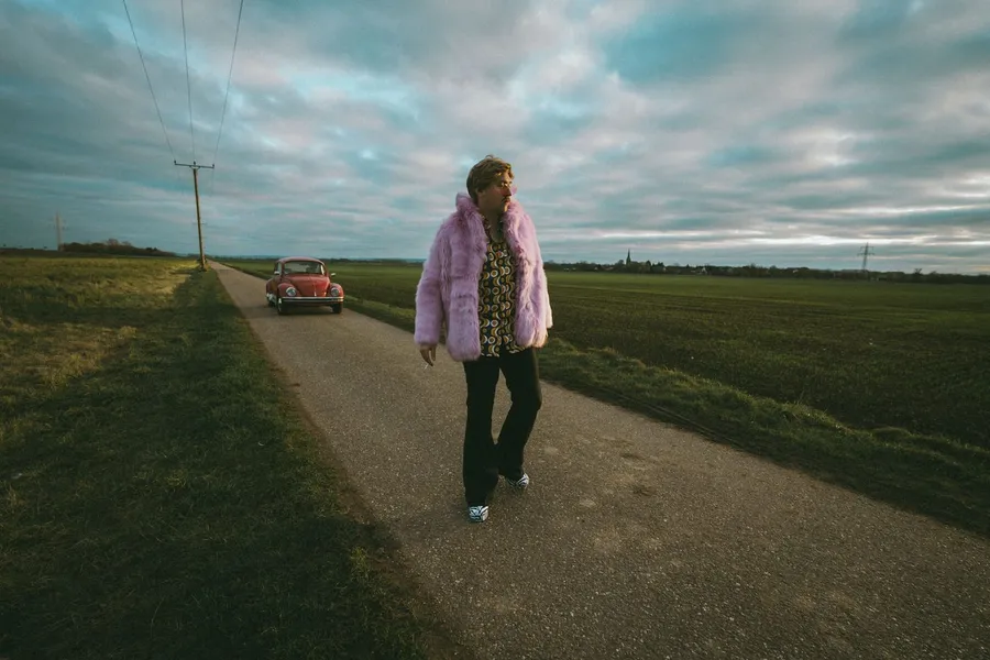 Person in a pink fur coat on a country road with a vintage Beetle and power lines, flash by Kilian Amrehn
