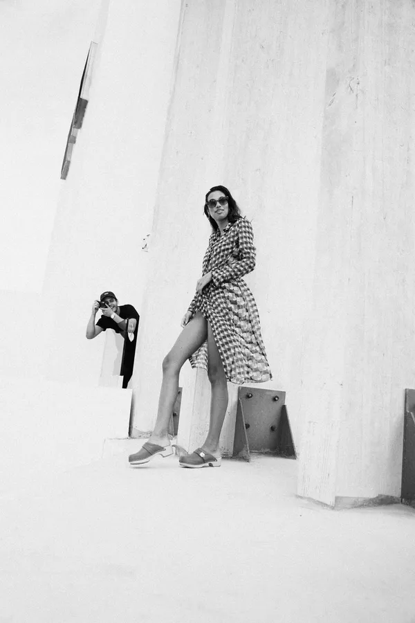 Woman in a coat poses by a white concrete wall, photographer in the corner, black and white, flash by Kilian Amrehn