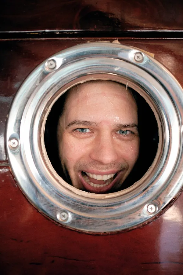 Grinning face of a man framed in a round metal porthole, flash by Kilian Amrehn