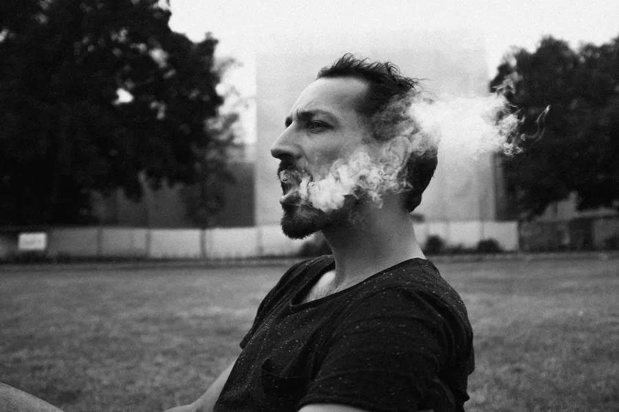 Man exhales smoke from mouth and nose in a park, black and white, flash by Kilian Amrehn