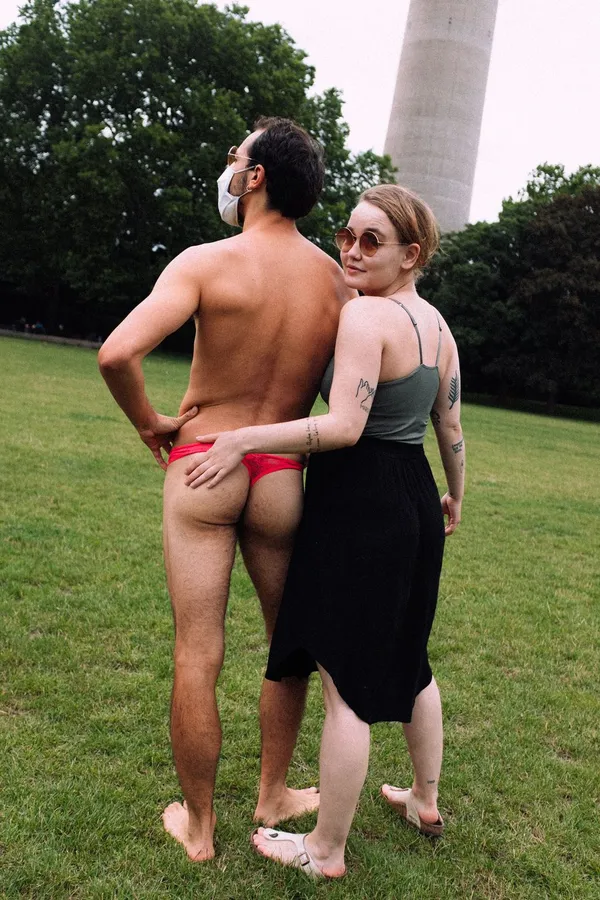 Man in a red thong and a woman embrace on a lawn, a tower in the background, flash by Kilian Amrehn