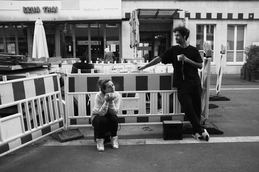 Two people with coffee at construction barriers outside a Thai restaurant, black and white, flash by Kilian Amrehn