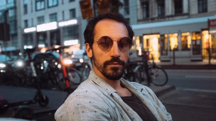 Bearded man in round sunglasses on an evening street lined with bicycles, flash by Kilian Amrehn