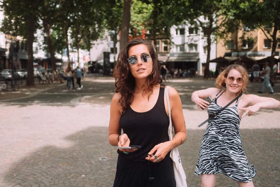 Two women on a sunny tree-lined square, one posing playfully in a zebra dress, flash by Kilian Amrehn