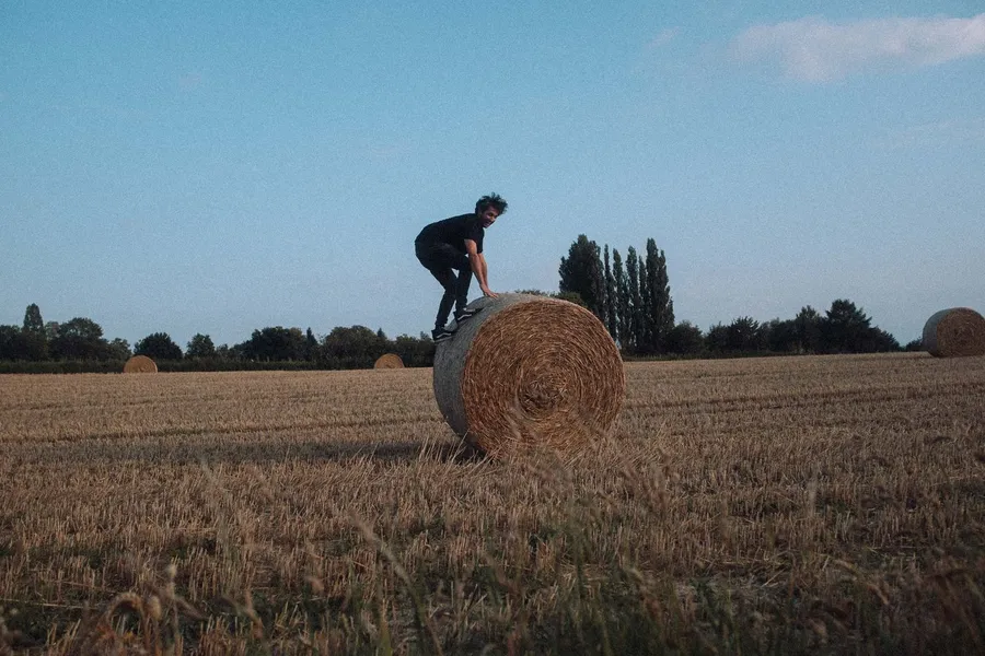 Man climbs onto a hay bale in a harvested field at dusk, flash by Kilian Amrehn