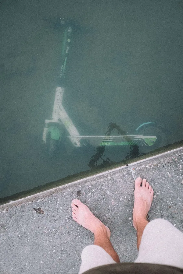 Bare feet at an edge above an e-scooter lying in murky water, flash by Kilian Amrehn
