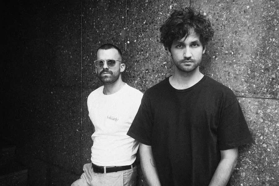 Two men in T-shirts lean against a granite wall, black and white, flash by Kilian Amrehn