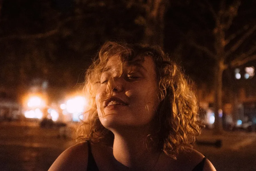 Curly-haired woman with closed eyes enjoys warm light at night, flash by Kilian Amrehn