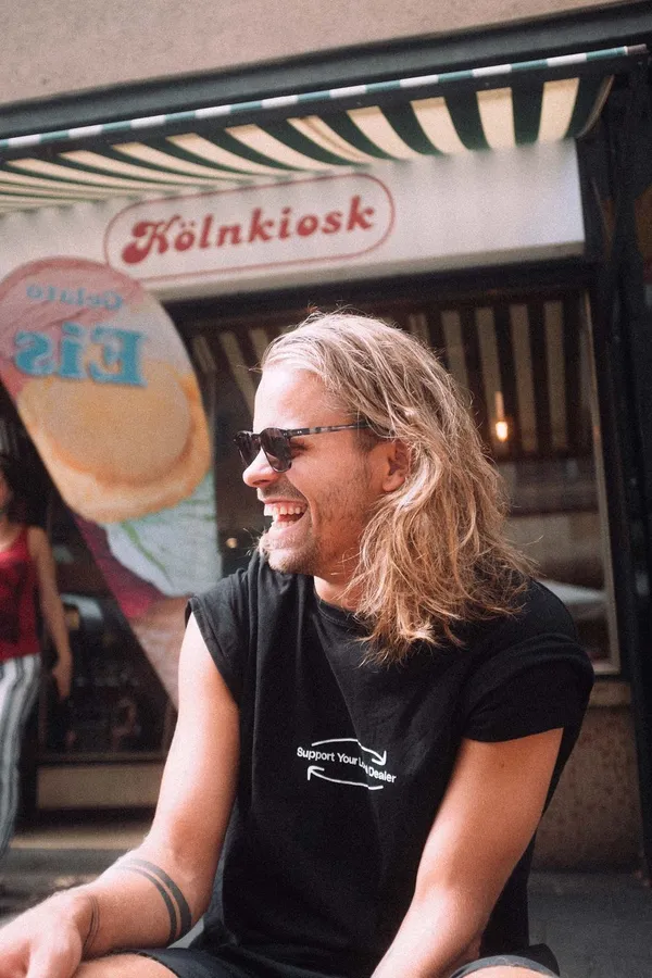 Laughing man with long blond hair and sunglasses in front of a kiosk, flash by Kilian Amrehn