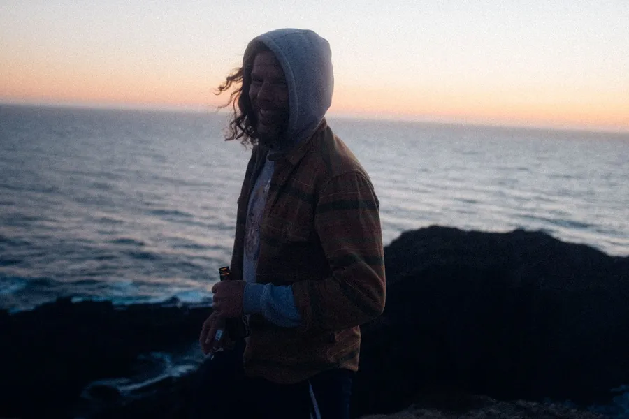 Laughing man in a hood standing on coastal rocks at sunset, Denmark, flash by Kilian Amrehn