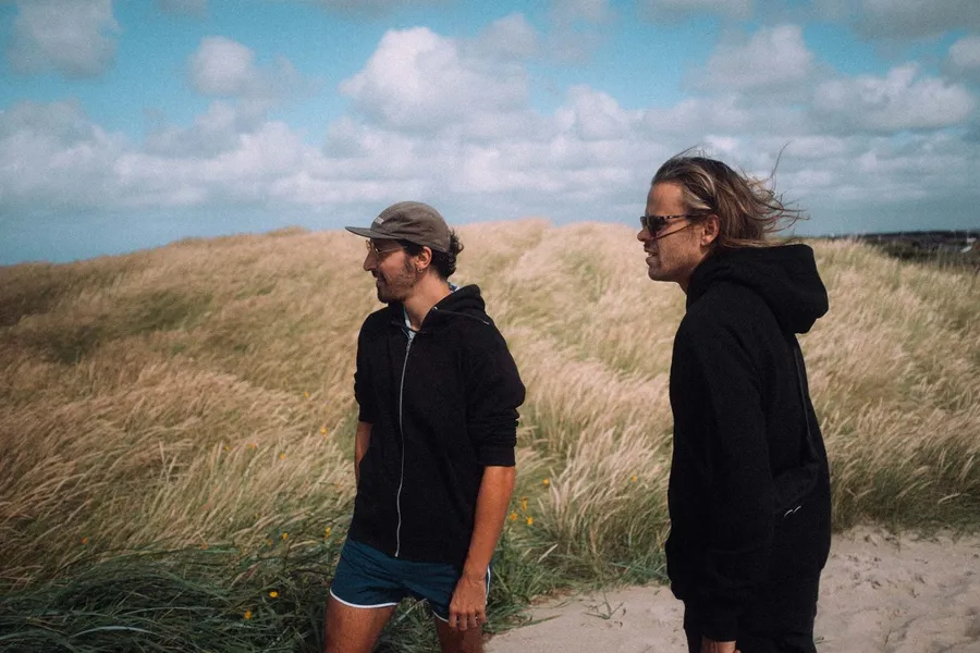 Two men in black hoodies among windswept grass dunes, Denmark, flash by Kilian Amrehn