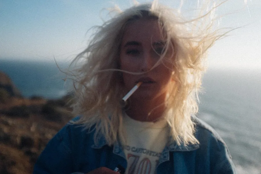 Blonde woman with a cigarette and windblown hair on the coast, Denmark, flash by Kilian Amrehn