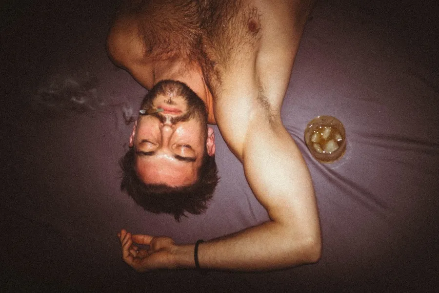 Shirtless man lies upside down on a bed beside a whisky glass, flash lit, Denmark, flash by Kilian Amrehn
