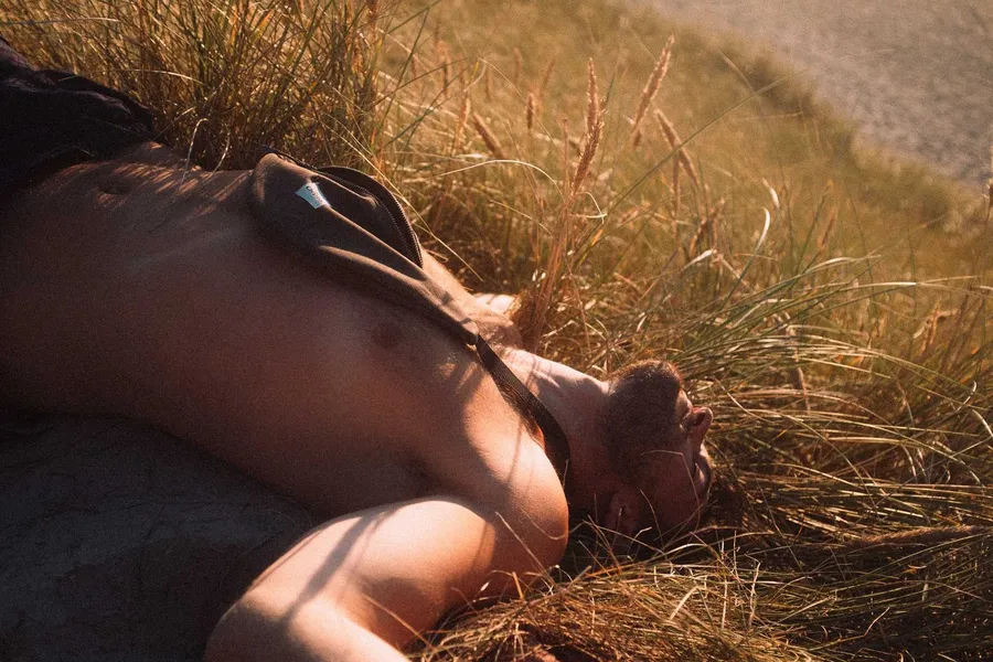 Shirtless man lies with eyes closed in sunlit dune grass, Denmark, flash by Kilian Amrehn