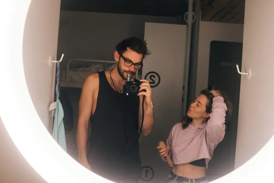 Mirror selfie of a man with a camera, a woman in a pink hoodie beside him, flash by Kilian Amrehn