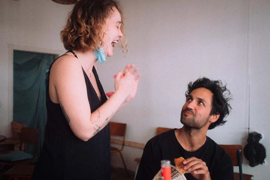 Laughing woman with a mask on her chin beside a man with a snack at a table, flash by Kilian Amrehn
