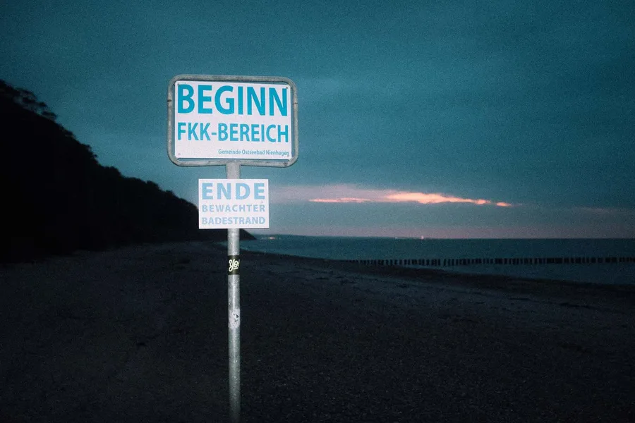 Sign reading Beginn FKK-Bereich, nudist area starts, on a beach at dusk, flash by Kilian Amrehn