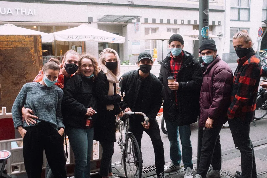 Group of friends with masks and coffee in front of a restaurant on the street, flash by Kilian Amrehn
