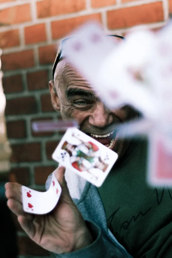 Laughing older man flings playing cards at the camera before a brick wall, flash by Kilian Amrehn