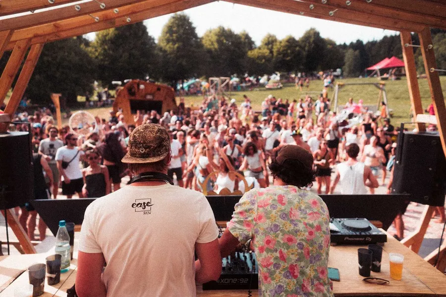 Two DJs seen from behind at the decks, dancing crowd on a festival field ahead, flash by Kilian Amrehn