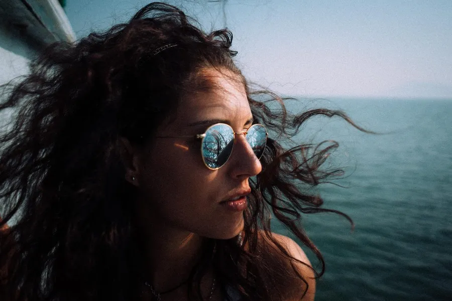 Woman in mirrored sunglasses with windswept curls above the sea, flash by Kilian Amrehn