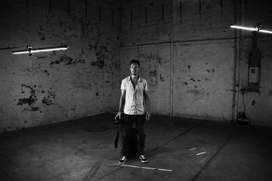 Man with a camera stands in an empty industrial hall with neon tubes, black and white, flash by Kilian Amrehn