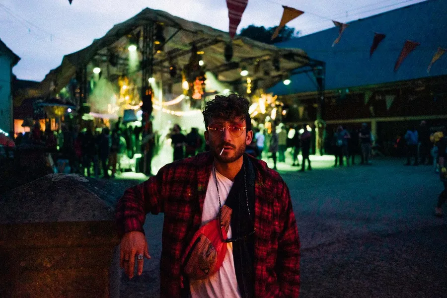 Man in a plaid shirt with a cigarette before a lit festival stage at dusk, flash by Kilian Amrehn
