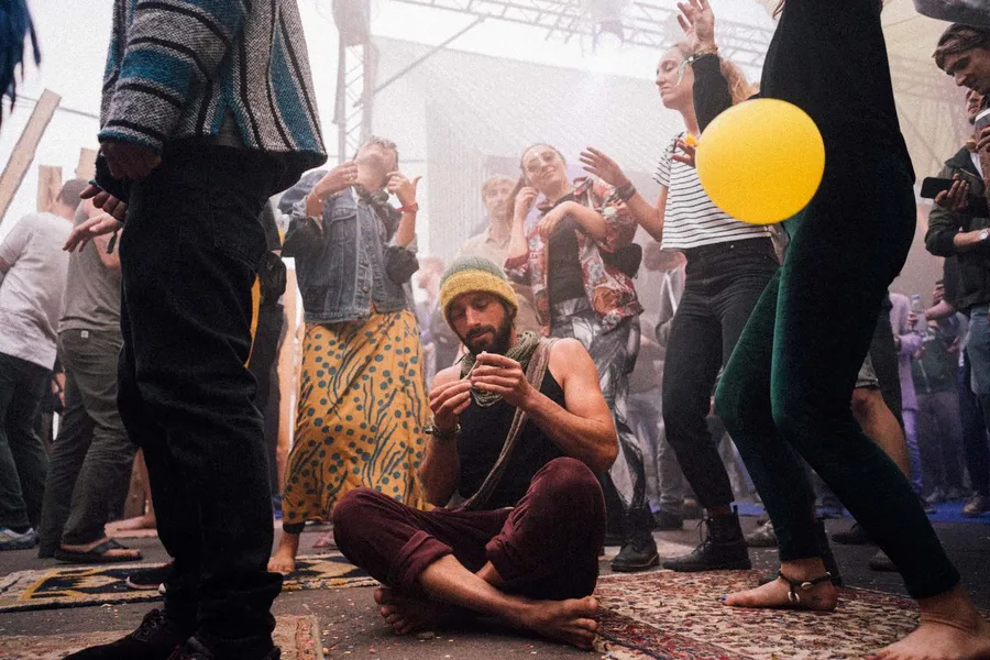 Cross-legged man on a rug amid a dancing crowd with a yellow balloon, flash by Kilian Amrehn