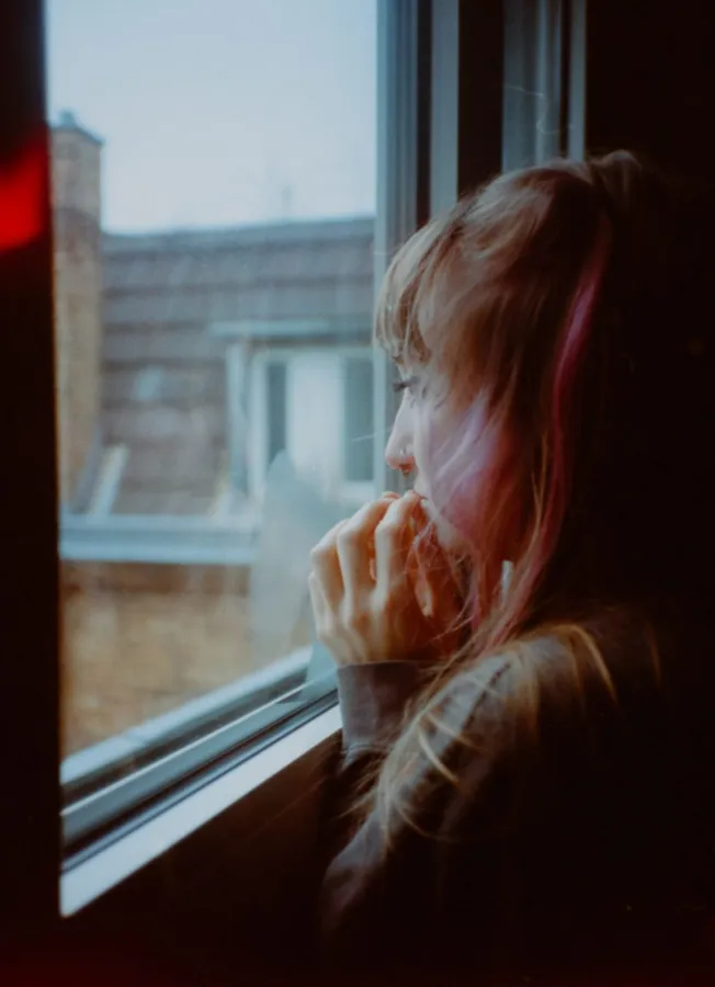 Woman with her hand to her mouth gazes out of a window at rooftops, analogue, flash by Kilian Amrehn