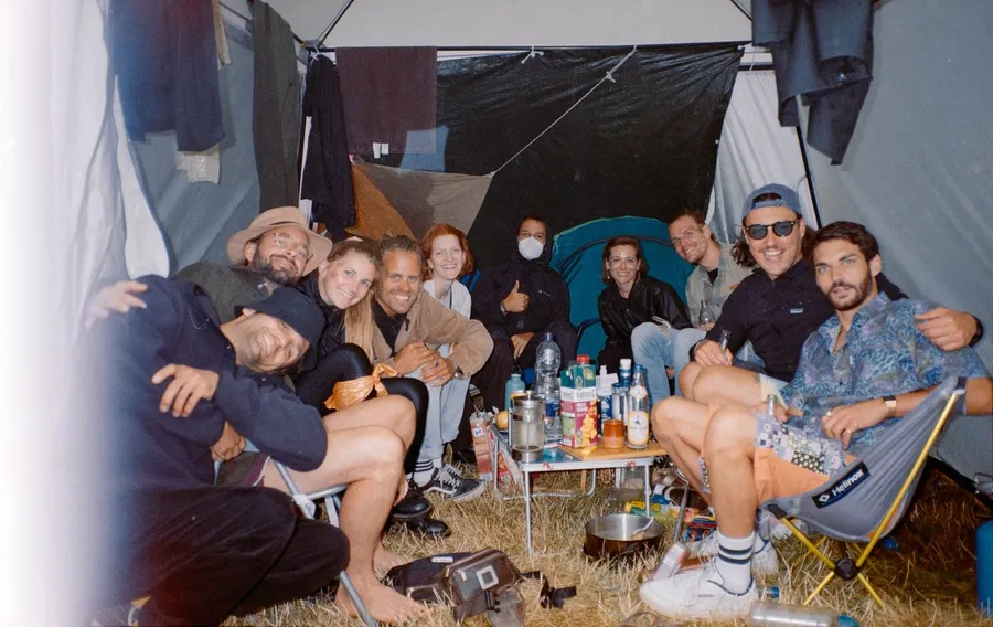 Cheerful group sits in a tent around a camping table with drinks, analogue, flash by Kilian Amrehn