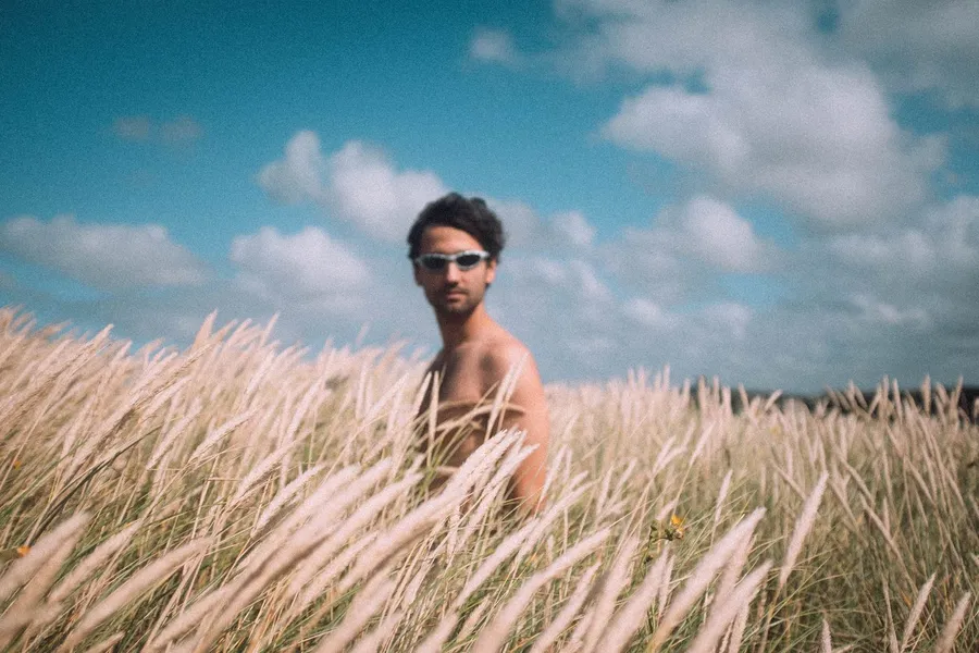 Man in sunglasses stands in tall marram grass under a cloudy sky, Denmark, flash by Kilian Amrehn