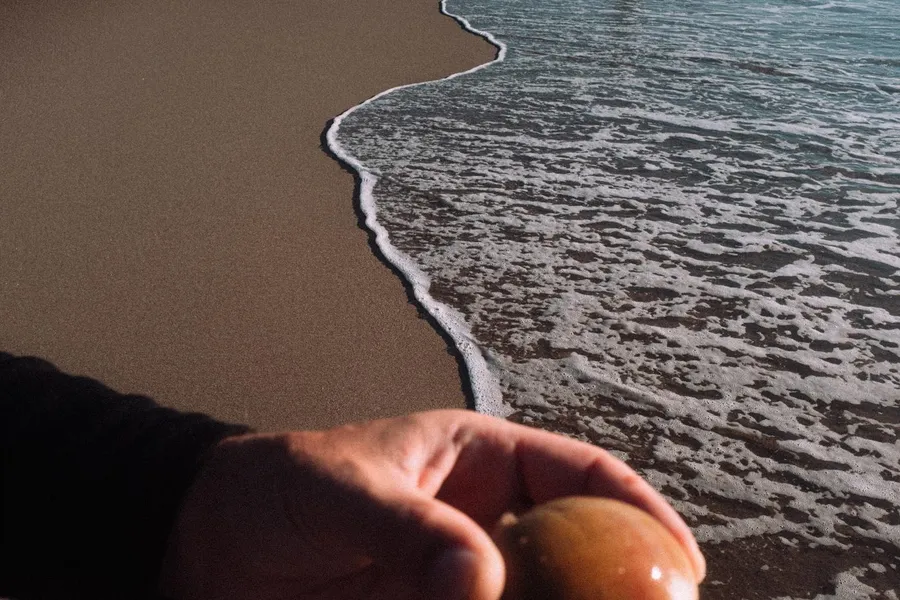 Hand holds an egg at the water's edge of a sandy beach, Denmark, flash by Kilian Amrehn