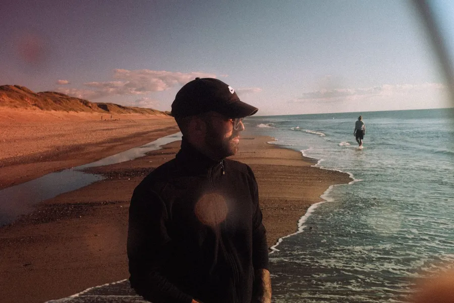Man in a cap on the beach in evening light, someone in the waves behind, Denmark, flash by Kilian Amrehn