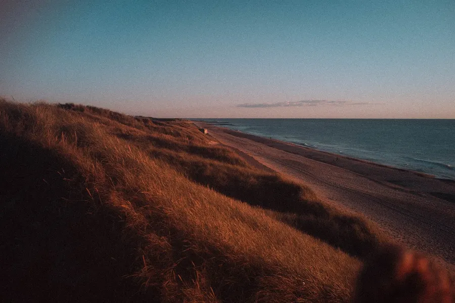 Grassy dunes in warm evening light above a long empty beach, Denmark, flash by Kilian Amrehn