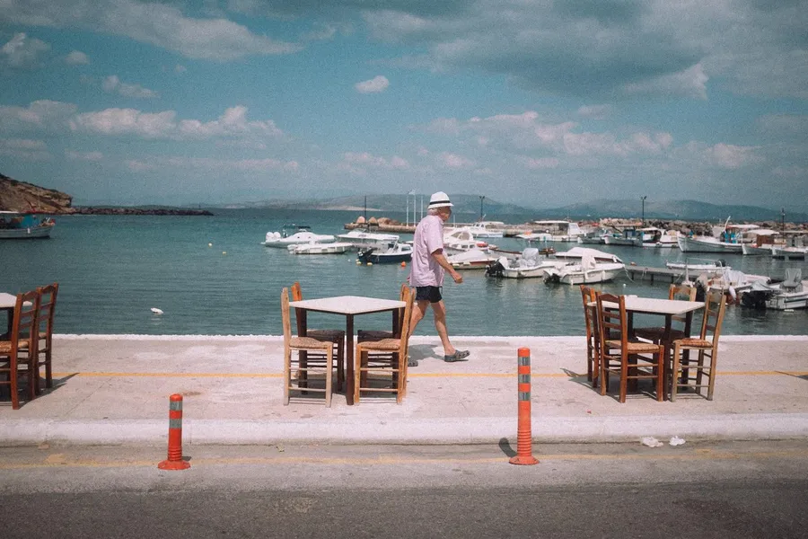 Man in a hat walks past taverna tables by a harbour full of boats, Greece, flash by Kilian Amrehn