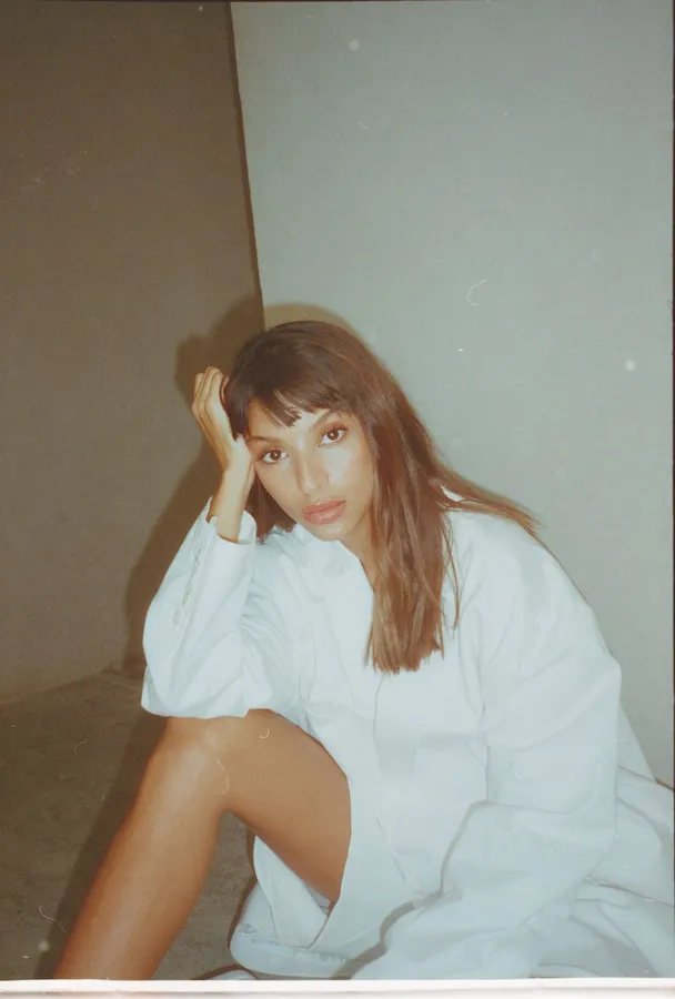 Woman in a white shirt sits in a room corner, head resting on her hand, analogue, flash by Kilian Amrehn