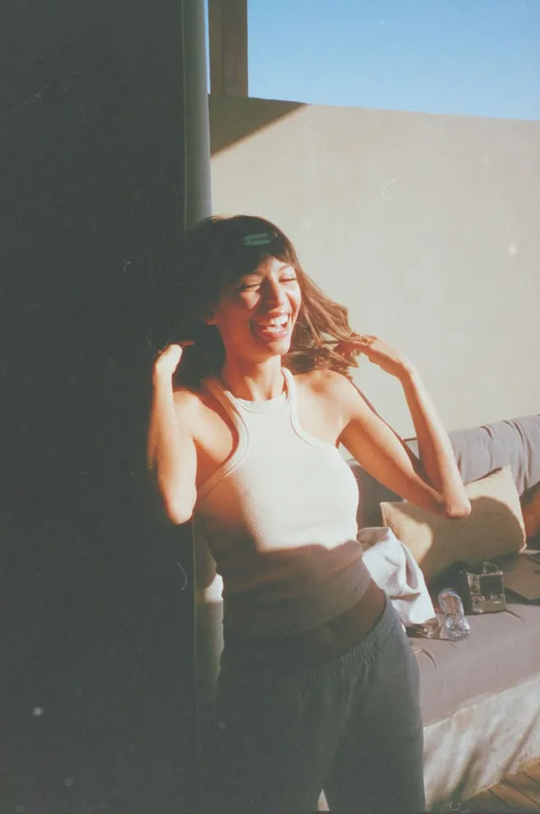 Laughing woman in a tank top in warm sunlight against a wall, analogue, flash by Kilian Amrehn