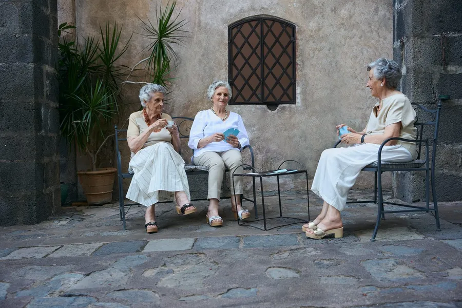 Three older women in white chat on benches in a stone courtyard, Scholl shoot, flash by Kilian Amrehn