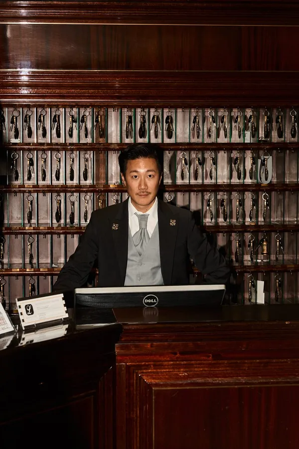 Man in a suit at a hotel reception desk before a wall of shoe rows, flash by Kilian Amrehn