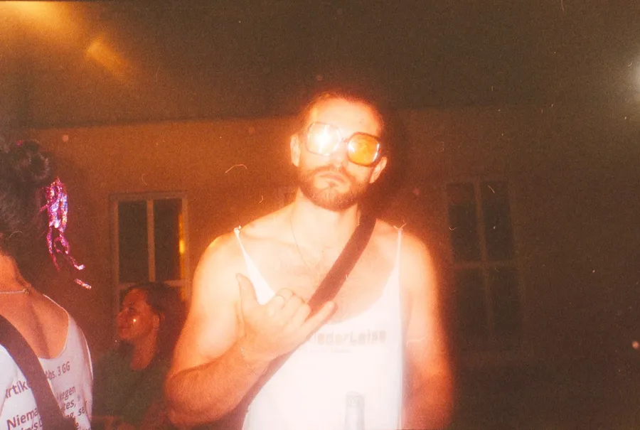 Man in tinted glasses making a shaka sign in warm party light, analogue, flash by Kilian Amrehn