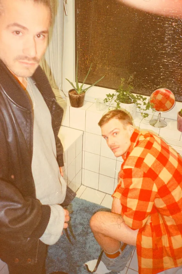 Two men in a tiled bathroom, one in a red check shirt looks up, analogue, flash by Kilian Amrehn