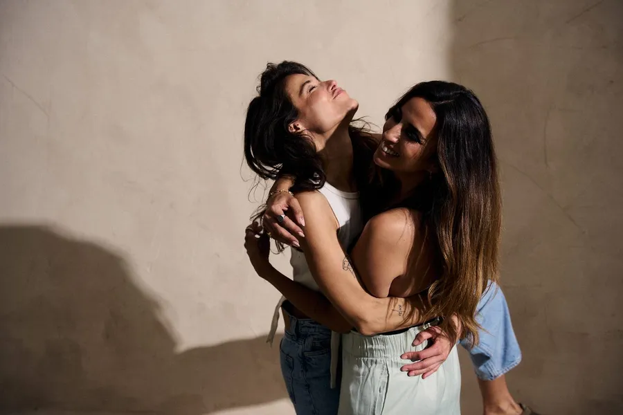 Two women embracing and laughing in front of a beige wall, hard shadow in the studio, flash by Kilian Amrehn