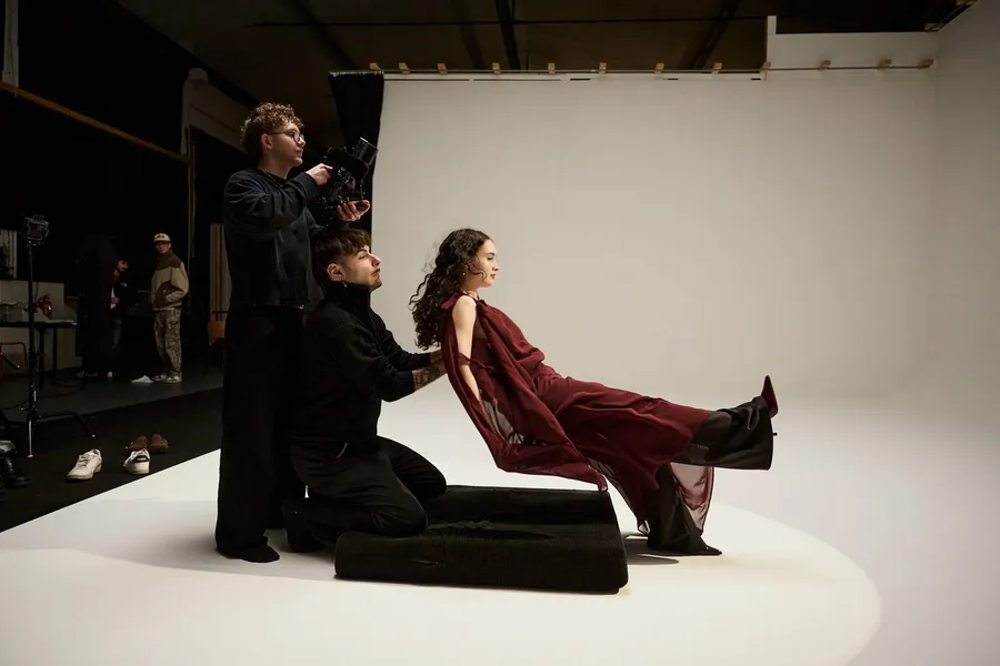 Backstage in a white studio: cameraman filming, woman in a dark red dress reclining on black cushions, flash by Kilian Amrehn