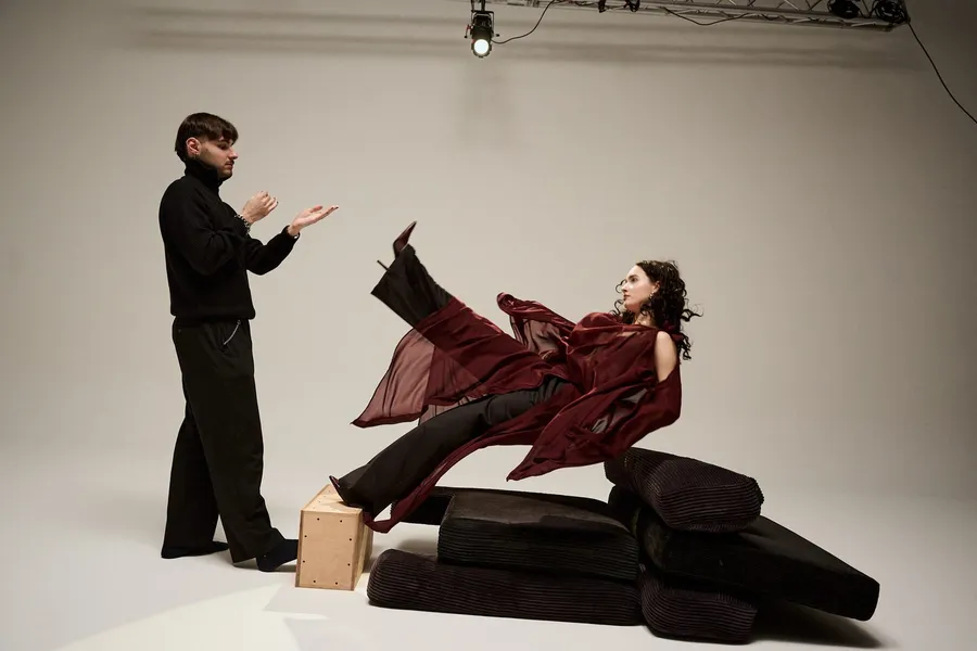 Man in black directing a woman in a flowing dark red dress on black cushions, white cyclorama, flash by Kilian Amrehn